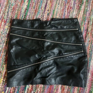 Svart skinnkjol från Mango - Säljer en supercool svart skinnkjol från Mango. Den är en del av deras Genuine Leather Collection och har en edgy look med flera dragkedjor som detaljer. Perfekt för en utekväll eller när du vill va cool i höst . Storlek EUR 34