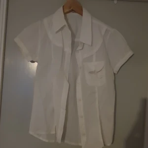 Vit kortärmad skjorta med knappar - Säljer en klassisk vit skjorta med korta ärmar och knappar framtill. Skjortan har en bröstficka med en liten rosett som detalj. Passar både till vardags och mer formella tillfällen.