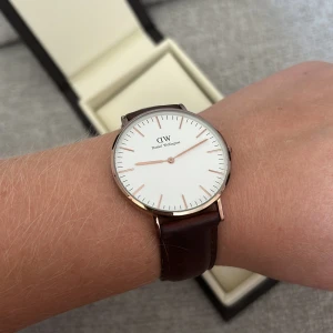 DW Klocka - Daniel Wellington klocka i guld med brunt läderband, tygband i marinblå och rosa medföljer. Jättefint skick, lite slitet på läderbandet o under uret, kolla bild