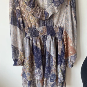 Mönstrad klänning från Zara - Säljer en superfin mönstrad klänning från Zara. Den har ett bohemiskt mönster i blå, beige och bruna toner. Klänningen är långärmad med volangdetaljer vid halsen och en smockad midja som ger en fin passform. Perfekt för både vardag och fest!