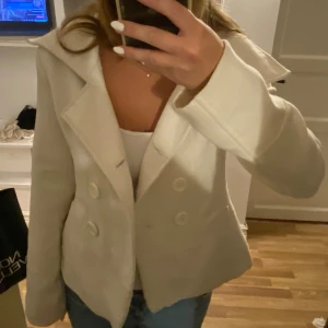 Vit kavaj med knappar - Säljer en snygg vit kavaj med stora knappar framtill. Den har en klassisk krage och är perfekt för både vardag och fest. Kavajen är långärmad och har en stilren design som passar till det mesta. Perfekt för att ge din outfit en elegant touch!