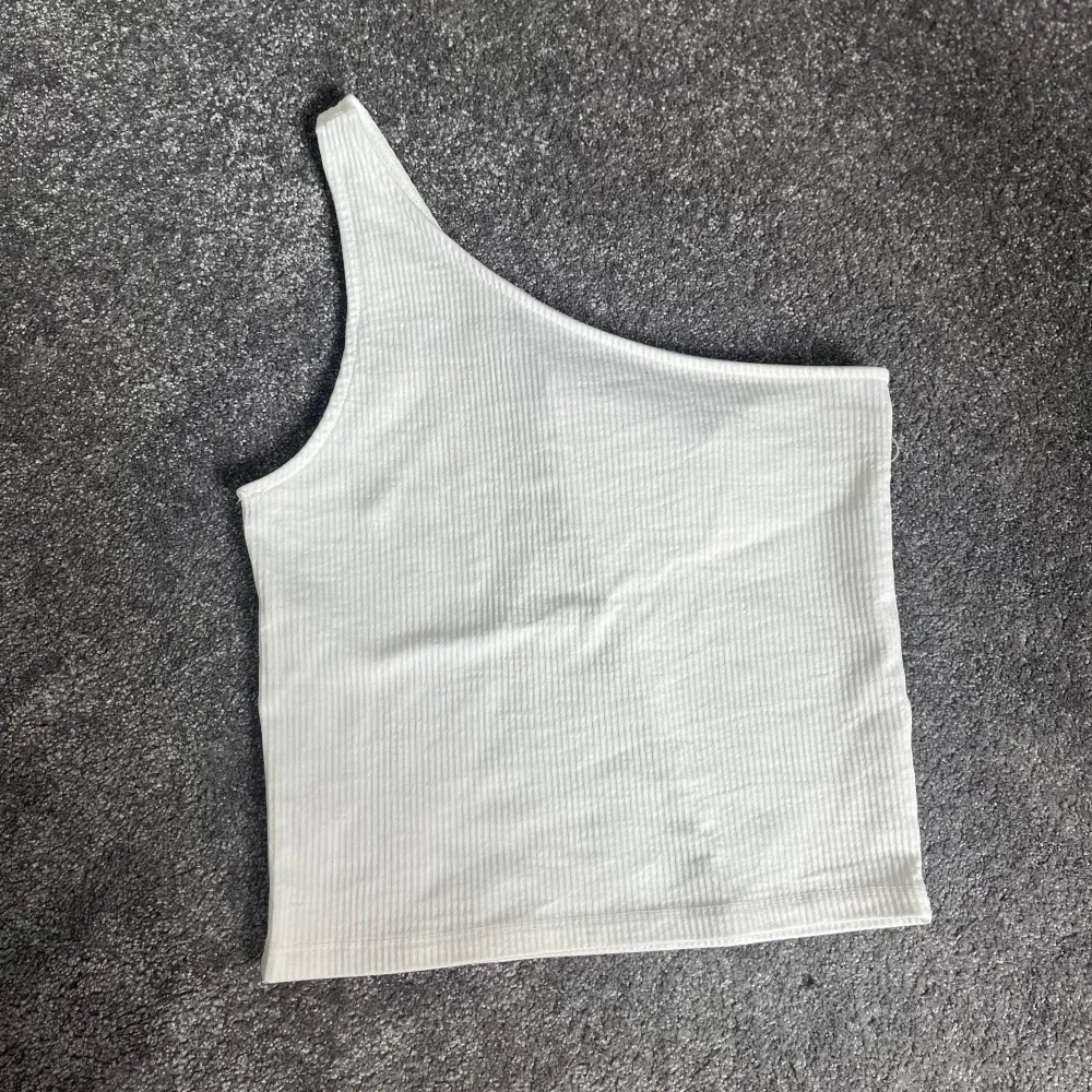 Säljer en stilren vit ribbad topp med oneshoulder-design. Perfekt för sommardagar eller en utekväll. Toppen är i ett mjukt och stretchigt material som sitter skönt på kroppen.. Toppar.