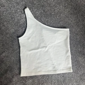 Vit ribbad oneshoulder topp - Säljer en stilren vit ribbad topp med oneshoulder-design. Perfekt för sommardagar eller en utekväll. Toppen är i ett mjukt och stretchigt material som sitter skönt på kroppen.