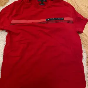 Säljer en snygg röd t-shirt från Tommy Hilfiger. Den har en cool randig detalj över bröstet med märkesloggan. Perfekt för en avslappnad stil och passar till både jeans och shorts. Skön och bekväm att ha på sig.