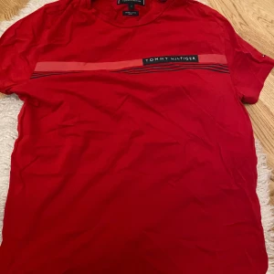 Röd t-shirt från Tommy Hilfiger - Säljer en snygg röd t-shirt från Tommy Hilfiger. Den har en cool randig detalj över bröstet med märkesloggan. Perfekt för en avslappnad stil och passar till både jeans och shorts. Skön och bekväm att ha på sig.