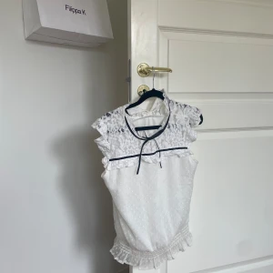 Vit spetsblus - Säljer en superfin vit blus. Den har vackra spetsdetaljer på axlarna och runt halsen, med en söt liten rosett framtill. Blusen är ärmlös och har en volangkant längst ner. Perfekt för sommarens fester eller en dag på stan!