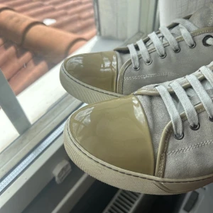 Beige sneakers med glansig tå - Säljer ett par snygga beige lanvins med en glansig Captoe! Skorna har snörning och en stilren design som passar perfekt till vardags. De är i bra skick och redo för nya äventyr!