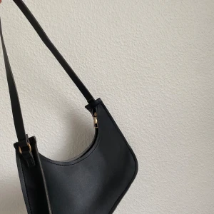 Svart axelväska i syntetmaterial - Säljer en stilren svart axelväska i syntetmaterial. Den har en enkel design med justerbar axelrem och guldfärgade detaljer. Perfekt för vardagsbruk eller en kväll ute. Väskan är i nyskick och redo för en ny ägare!