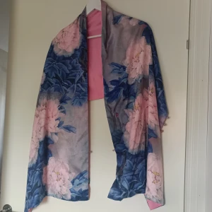 Blommig sjal med rosa och blå detaljer - Säljer en superfin sjal med ett blommigt mönster i rosa och blått. Den har en mjuk och elegant känsla, perfekt för att piffa upp vilken outfit som helst. Sjalen har en ljusrosa innerfoder och  små knappar som ger en extra touch av stil. Perfekt för sensommar och höst!
