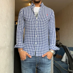 Blårutig skjorta från Ralph Lauren - Säljer en snygg blårutig skjorta från Ralph Lauren. Skjortan har långa ärmar och är i ett klassiskt rutigt mönster med vita och blå rutor. Den har knappar framtill och en liten röd broderad logga på bröstet. Perfekt för både vardag och fest!