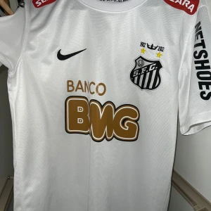 Vit fotbollströja från Nike - Säljer en vit fotbollströja från Nike med S.F.C.-logga och sponsorer som Banco BMG och Netshoes. Tröjan har korta ärmar och är tillverkad i Dri-FIT-material för att hålla dig sval och torr. Perfekt för fotbollsfans!
