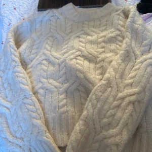 Beige stickad tröja - Mysig beige stickad tröja med flätmönster. Perfekt för kyliga dagar med sin varma och bekväma design. Tröjan har långa ärmar och en rund halsringning.