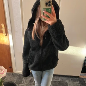 Svart teddyjacka med dragkedja - Mysig svart teddyjacka perfekt för kyliga dagar. Den har en dragkedja framtill och långa ärmar. Jackan är i ett mjukt och fluffigt material som håller dig varm och bekväm. Perfekt för höst och vinter!