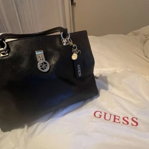 Svart handväska från Guess - Snygg svart handväska från Guess med silverdetaljer och logotypmönster. Väskan har både handtag och en avtagbar axelrem i kedja. Den har en dragkedja på baksidan och flera fack inuti för praktisk förvaring. Oanvänd❤️❤️❤️