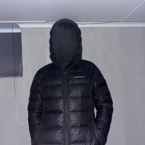 Svart dunjacka från Peak Performance - Säljer en svart dunjacka från Peak Performance. Jackan har en dragkedja framtill och en huva för extra skydd mot kyla. Perfekt för kalla dagar. Som ni kan se så har den ett litet hål som läker dun ibland men inte alls ofta. 