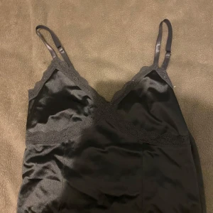 Grå satinlinne med spetsdetaljer - Elegant grå linne i satin med smala axelband och spetsdetaljer längs halsringningen. Perfekt för en stilren och sofistikerad look.