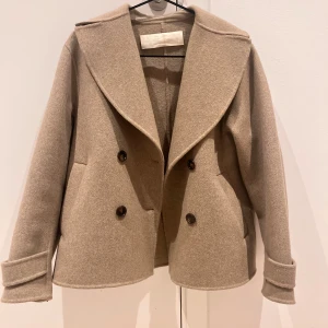 Beige kappa från Arket - Säljer en stilren beige kappa från Arket. Jackan har en dubbelknäppning och breda slag. Perfekt för höst och vår, med långa ärmar och en elegant look. Den är väldigt ovetandes 