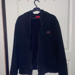 Hugo Boss Zip Up Hoodie - Snygg svart zip hoodie från Hugo med dragkedja och huva. Den har en diskret logga på bröstet och långa ärmar. Perfekt för en avslappnad stil. 1600kr nypris på Zalando. Slutsåld finns inte mer. Pris kan diskuteras.