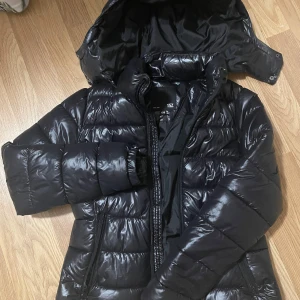 Svart pufferjacka från 157 - Snygg svart pufferjacka från 157 med glansig finish och huva. Jackan har dragkedja framtill och är perfekt för kyligare dagar. Den är i storlek dam xxs och har långa ärmar för extra värme.