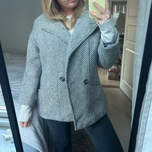 Grå randig kappa - Säljer en stilren grå randig kappa med knappar framtill. Jackan har en klassisk krage och långa ärmar, perfekt för kyligare dagar. Passar bra till både jeans och mer uppklädda outfits.