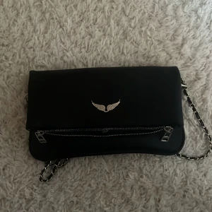 Svart axelväska med kedja och vingdetalj - Snygg svart axelväska med en elegant kedjerem och en cool vingdetalj på framsidan. Väskan har en praktisk dragkedja och magnetknappar för enkel öppning och stängning. Perfekt för att ge din outfit en stilfull touch.
