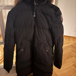 Svart dunjacka från Parajumpers - Säljer en svart dunjacka från Parajumpers med pälsfodrad huva och dragkedja. Jackan har långa ärmar och praktiska fickor med knappar. Perfekt för kalla vinterdagar.