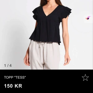 Svart topp 'Tess' - Säljer en svart topp med v-ringning och volangdetaljer. Den har korta ärmar och en luftig passform, perfekt för både vardag och fest. Toppen är i ett mjukt och bekvämt material som faller fint.