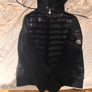 Moncler Cardigan - Säljer en snygg svart jacka/cardigan från Moncler. Den har en pufferdesign på framsidan och ärmarna, med en dragkedja och huva. Perfekt för kyliga dagar och ger en stilren look. Passar både till vardags och lite finare tillfällen. Sitter som storlek 1 / S