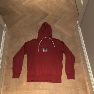 Röd hoodie från Gant - Säljer en snygg röd hoodie från Gant. Använd och har fått lite skada vid snörena, därmed lägre pris