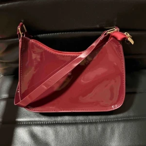 Vinröd axelväska med dragkedja - Snygg vinröd axelväska i glansigt material med dragkedja upptill! Mått: 23x17 cm, aldrig använd