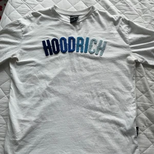 Vit t-shirt från Hoodrich - Säljer en snygg vit t-shirt från Hoodrich i storlek S. Den har ett coolt broderat märke i blå nyanser på framsidan. Perfekt för en avslappnad stil eller att bära till vardags. T-shirten är i bra skick och gjord av mjukt bomullsmaterial.