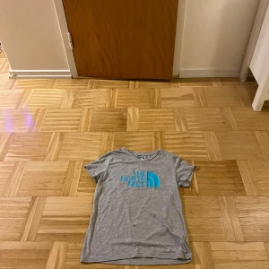 Grå t-shirt från The North Face (Barnstorlek) - Säljer en grå t-shirt från The North Face med turkos logga på bröstet. Perfekt för en avslappnad stil. T-shirten har korta ärmar och är tillverkad i ett mjukt material. Den har även en snygg V ring vid halsen och är storlek L i Barn vilket motsvarar ca 152-158
