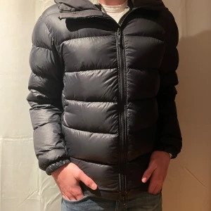 Svart dunjacka från J.Lindeberg - Säljer en svart dunjacka från J.Lindeberg i storlek L. Jackan är i mycket bra skick och perfekt för kalla vinterdagar. Den har en dragkedja framtill och en mysig huva. Passar både till vardags och lite finare tillfällen. Håll dig varm och stilren i vinter! 