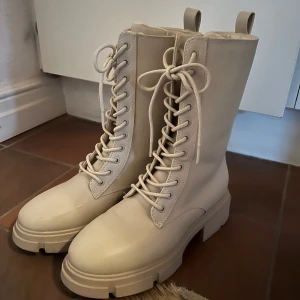 Beiga  boots med snörning strl 39 - Snygga beiga boots med snörning och grov sula. Perfekta för en stilren look med en touch av edge. De har en hög skaft och är tillverkade i syntetmaterial. Storlek 39 ifrån New Yorker, knappt använda. (Lite smutsiga framtill bara).