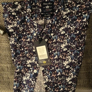 Helt ny Blommig skjorta från Robbie Moor - Snygg blommig skjorta från Robbie Moor i storlek XL. Skjortan har ett färgglatt mönster med inslag av blått, vitt och brunt. Perfekt för att liva upp garderoben med en touch av naturinspirerat mönster.