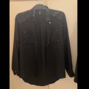 Svart skjorta med knappar - Snygg svart skjorta med långa ärmar och knappar framtill. Två bröstfickor med knappdetaljer ger en stilren look. Perfekt för en elegant och avslappnad stil.