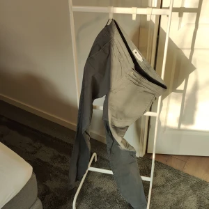Grå byxor från NN07 - Säljer ett par stilrena grå byxor från NN07 i storlek 30/32. De har en klassisk design med dragkedja och knappar, samt praktiska bakfickor. Perfekta för både vardag och mer uppklädda tillfällen. Passar bra till höst- och vårsäsongen.