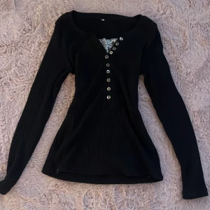 Svart långärmad tröja med knappar - Säljer en stilren svart tröja med långa ärmar och snygg v-ringning. Tröjan har dekorativa knappar framtill och en fin spetsdetalj vid halsen. Perfekt för downtown girls🖤