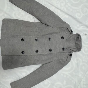 Grå kappa från H&M - Säljer en stilren grå kappa från H&M i storlek M. Den har en dubbelknäppt design med svarta knappar och långa ärmar. Perfekt för höst och vår, och ger en elegant touch till vilken outfit som helst. Passar både till vardags och lite finare tillfällen.