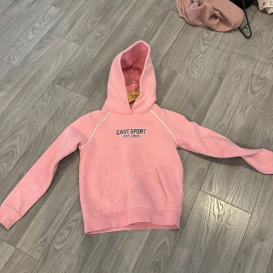 Rosa hoodie från GANT - Säljer en snygg rosa hoodie från GANT med texten 'GANT SPORT EST. 1949' på bröstet. Den har en stor ficka framtill och en bekväm huva med GANT-logga. Perfekt för en avslappnad stil.