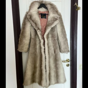 Beige kappa i fuskpäls - Elegant och lyxig beige kappa i fuskpäls med mjukt foder i rosa. Perfekt för att hålla sig varm och stilfull under kyliga dagar. Köptes på plick för 200 kr förra året, men har inte kommit till användning då jag tycker den känns något liten för min smak. Jag är 172 cm lång och har storlek S. Det står ingen specifik storlek på jackan men den känns som en xs och skulle nog sitta bättre på någon som är lite kortare. Jackan saknar knappar för stängning, men utöver det känns den jättefin.