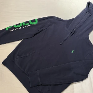 Hoodie från Ralph Lauren - Snygg mörkblå hoodie från Ralph Lauren med ett unikt grönt POLO-tryck på ärmen och en liten grön logga på bröstet. Perfekt för en avslappnad stil med sin klassiska design och bekväma passform.