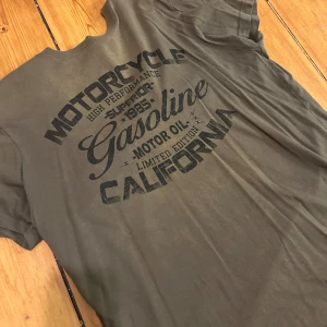 Olivgrön t-shirt från Bluewear Co. - Säljer en olivgrön t-shirt från Bluewear Co. med tryck på ryggen som säger 'Motorcycle High Performance Gasoline California'. Perfekt för en avslappnad stil.