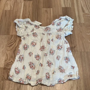 Blommig topp från Newbie - Söt blommig topp från Newbie i storlek 74 för 6-9 månader. Toppen har korta ärmar med spetsdetaljer och ett romantiskt blommönster i rosa och grönt på en beige bakgrund. Perfekt för varma dagar!