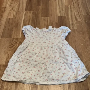 Blommig klänning från H&M - Söt blommig klänning från H&M i ljus färg med små rosa blommor. Klänningen har korta ärmar och en rund halsringning. Perfekt för små barn som vill känna sig fina.