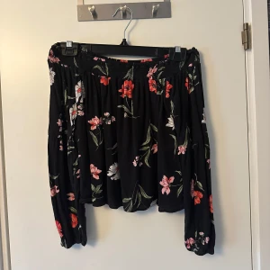 Svart blommig blus från Hollister - Säljer en svart blus från Hollister med ett vackert blommönster i rosa, rött och vitt. Blusen har långa ärmar och en luftig passform, perfekt för både vår och sommar. Blusen är även off shoulder