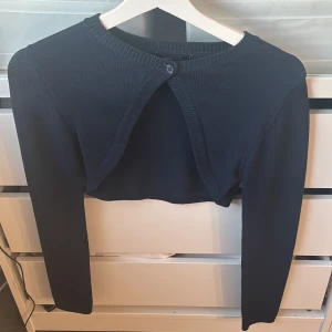 Mörkblå bolero - Säljer en snygg mörkblå ribbad bolerotopp med långärmad design. Perfekt för lager-på-lager-stil eller som ett extra lager under kyliga kvällar. Toppen är croppad och har en knapp framtill för en stilren look. Passar perfekt till både vardag och fest! Säljer pågrund av att den ej kommer till användning. Endast använd vid ett tillfälle!