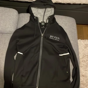 Svart hoodie från Hugo Boss - Säljer en svart hoodie från Hugo Boss i regular fit. Den har en dragkedja framtill och vita detaljer vid fickorna. Perfekt för höst och vår, och den är i mycket bra skick. Passar både till vardags och lite mer avslappnade tillfällen.