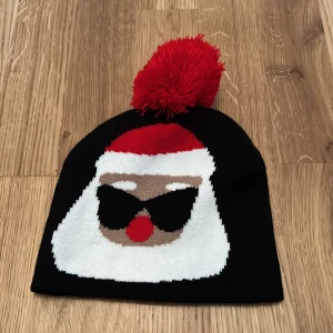 Svart mössa med tomtemotiv - Snygg svart mössa med ett roligt tomtemotiv framtill och en röd tofs på toppen. Perfekt för att sprida julstämning under vintern!