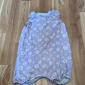 Ljuslila blommig klänning - Söt ljuslila klänning med blommigt mönster från I Love Eco. Tillverkad i ekologisk bomull, perfekt för en bekväm och stilren look. Klänningen har volangdetaljer vid halsen och ärmlös design.